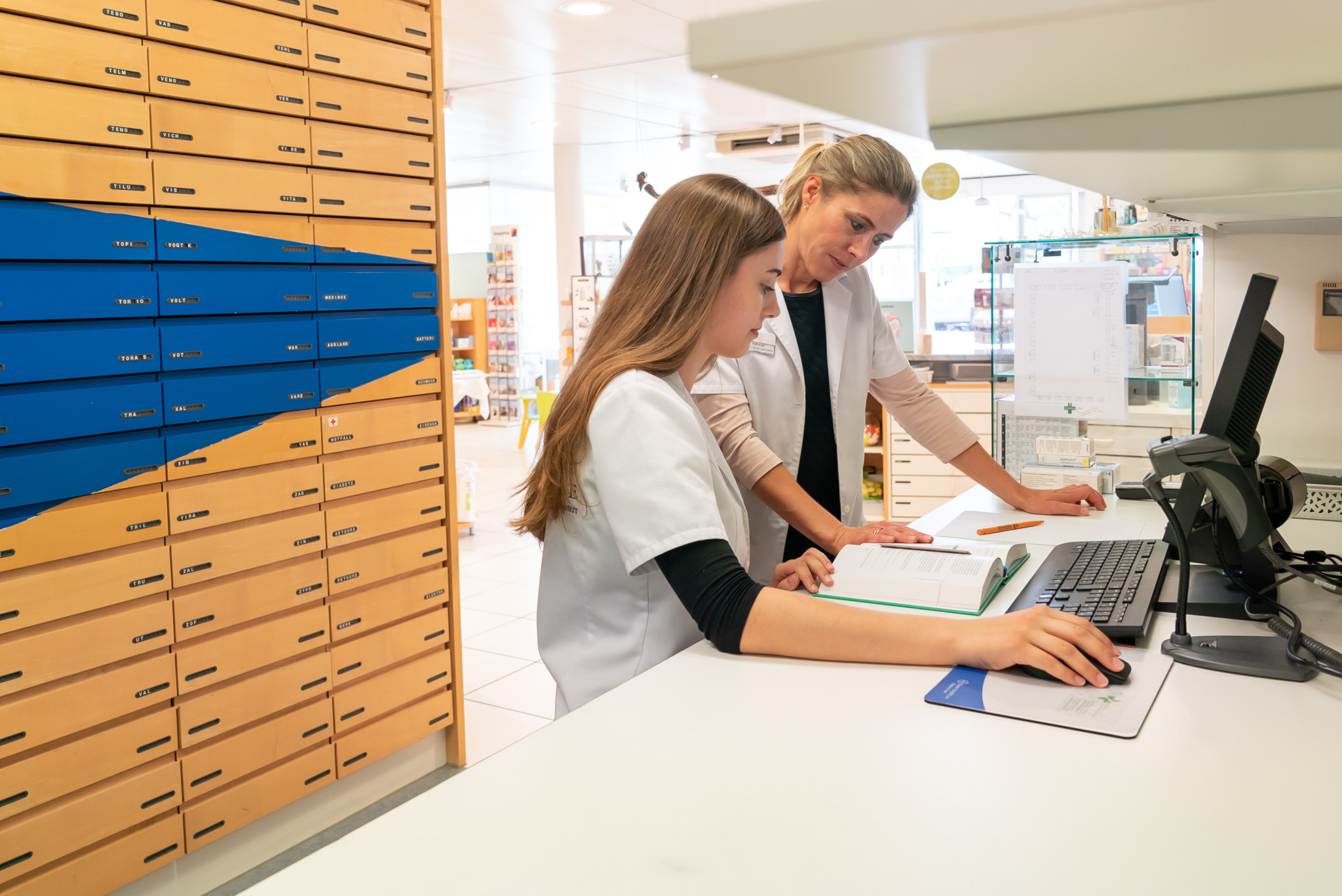 Une pharmacienne blonde aide une jeune apprentie à étudier des livres médicaux sur un bureau. Une pharmacienne blonde aide une jeune apprentie à étudier des livres médicaux sur un bureau.