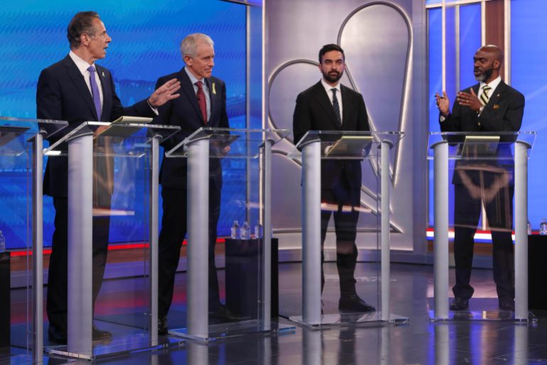 Andrew Cuomo, Michael Blake, Zohran Mamdani and Whitney Tilsen stand behind glass podiums at a debate