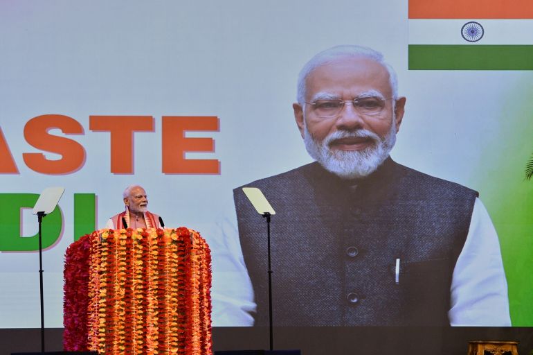 Narendra Modi speaks at a podium. Behind him is a screen projecting his picture and an Indian flag.