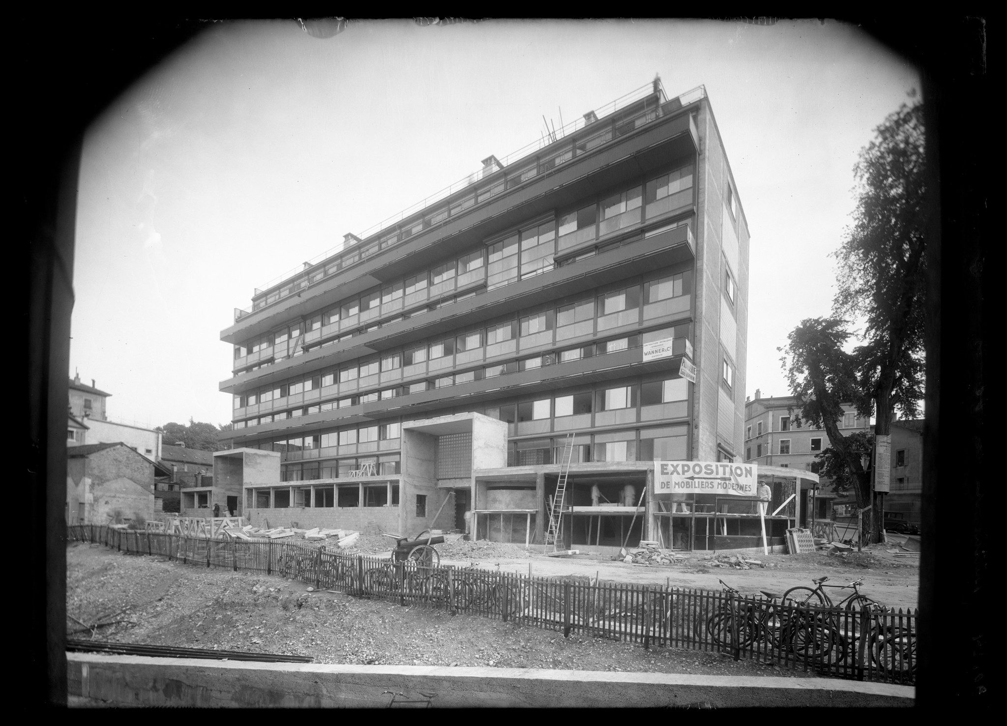 Bâtiment moderne en construction avec une grande pancarte ’EXPOSITION’, échafaudages et vélos stationnés à l’avant.