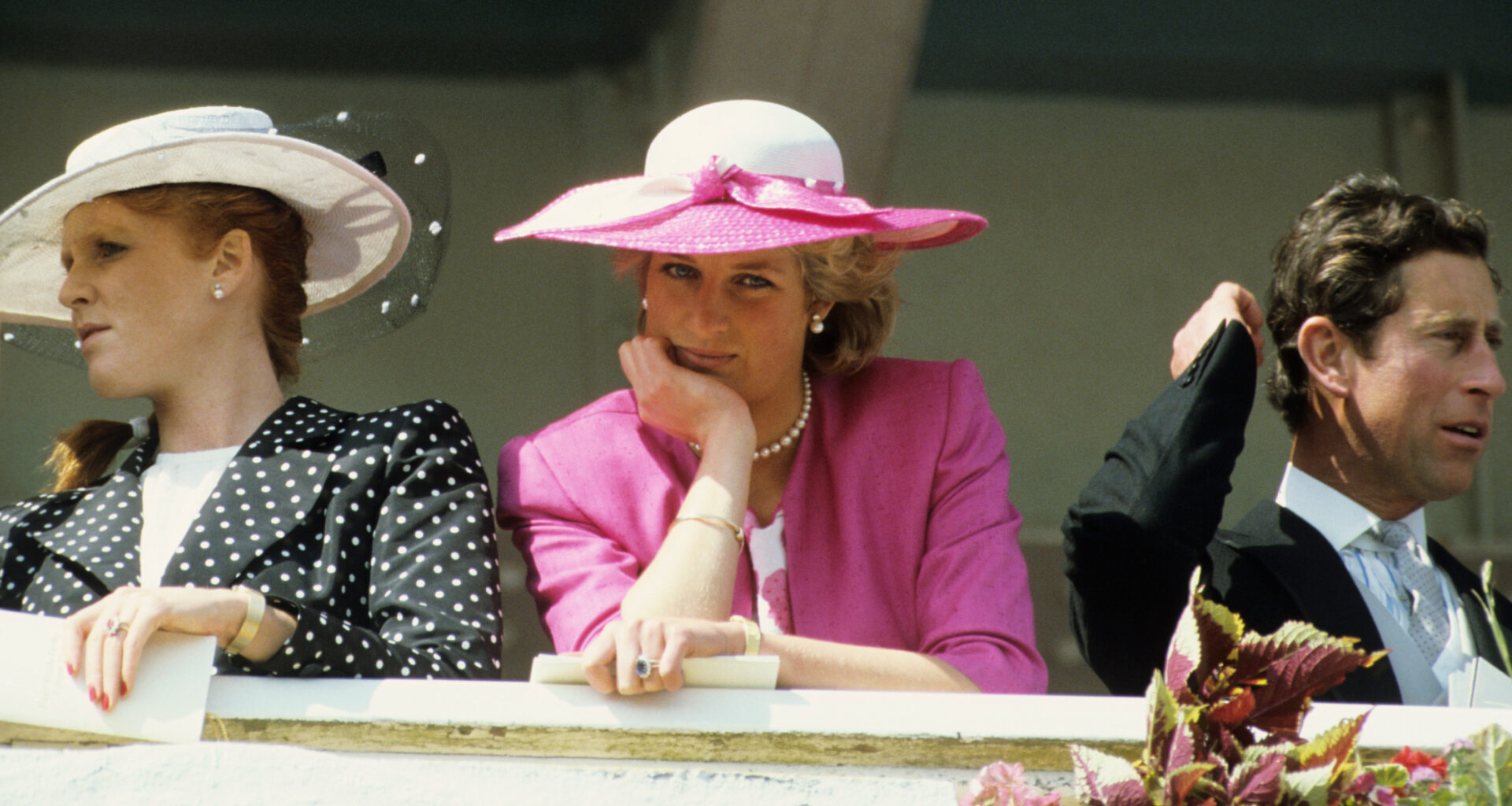 Princess Diana wearing a pink blazer and white hat resting her chin on her hand at the 1987 Epsom Derby next to Sarah Ferguson in a black polka dot coat and Prince Charles in a suit