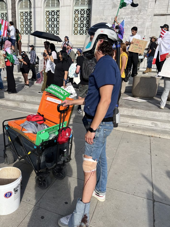 A medic with a full face mask stands with the left leg of their jeans rolled up past their knee. Blood drips down their leg from their knee, which is wrapped in gauze and bandage. They are holding onto an "eye-washing" mobile cart.