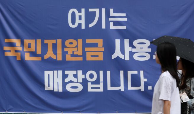 Ahead of the July 21 launch of President Lee Jae-myung’s “Consumer Coupon for Livelihood Recovery” program, optimism is growing among traditional markets, neighborhood grocers, and convenience store operators about a potential economic rebound. Aimed at stimulating domestic consumption, the government plans to distribute up to ₩550,000 in coupons per person over two phases. The photo shows a notice posted at a shop in Seoul indicating it as a designated location for using the consumer coupons. (Photo courtesy of Yonhap)