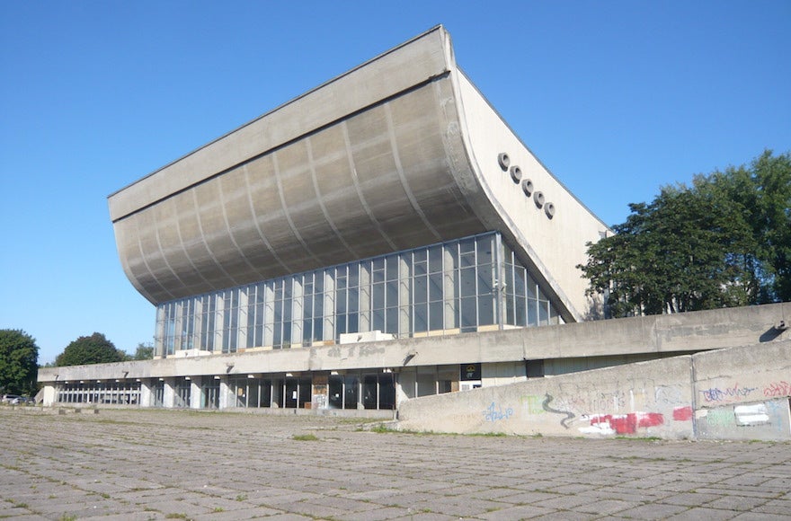 A photo of the Vilnius Palace of Concerts and Sports. It is an example of brutalist architecture and has a large roof that swoops upwards above a wall of windows.