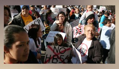 A photo shows a crowd of mostly Latino people of different ages holding signs and wearing shirts against HB-56.