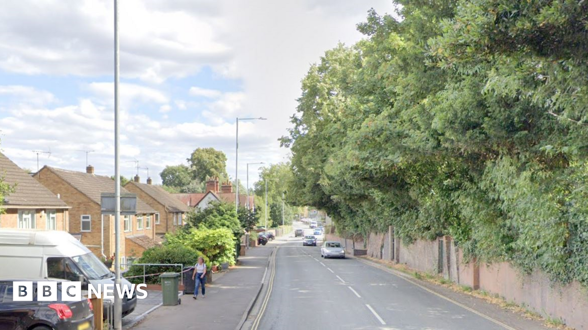 Hylton Road in Worcester with a wall on one side and houses on the other, with cars and vans parked outside. There are trees, ivy and shrubs overhanging the wall and there are cars driving in the road. A woman is walking on a pavement near a wheeled bin.