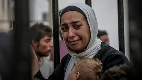 Getty Images A Palestinian woman wearing a headscarf cries with one hand holding a baby to her chest and another clutched to iron bars that she is stood next to.