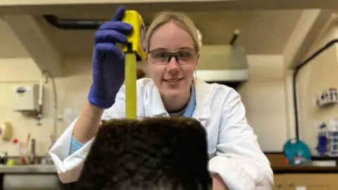 BBC Ellie is looking at the camera and is wearing protective glasses. She is sitting behind a lump of peat and holding a yellow tape measure beside it. She is wearing blue protective gloves and a white lab coat. She has blonde hair tied back.