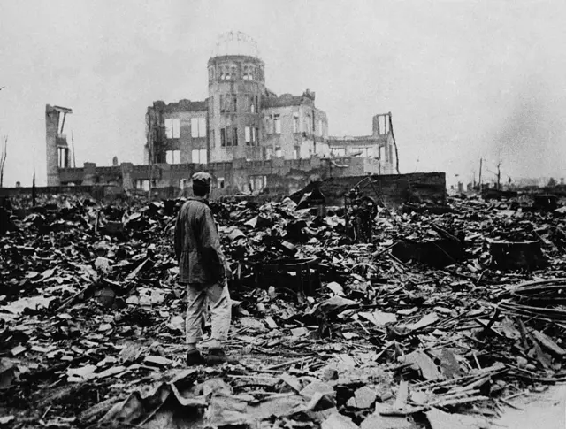 A man stand among di ruins of Hiroshima, wit di skeleton of a large building for front of am, an atomic bomb drop dia