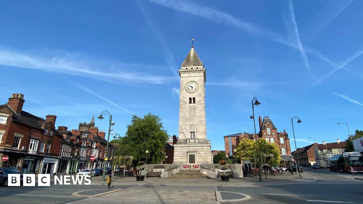 In the centre of the image of a town centre is a large light-coloured stone column memorial. It has a pointed slate roof and a clock as well as brass plaques. Near the plaques are red poppy wreaths. The memorial is on an island in the middle of a road - on either side are shops and cars.