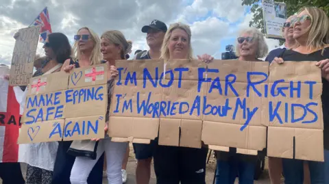 Greg McKenzie/BBC A line of women holding handmade cardboard banners, saying "Make Epping Safe Again" and "I'm not far right, I'm worried about my kids". Some are wearing sunglasses. The banners also bear hearts and England flags