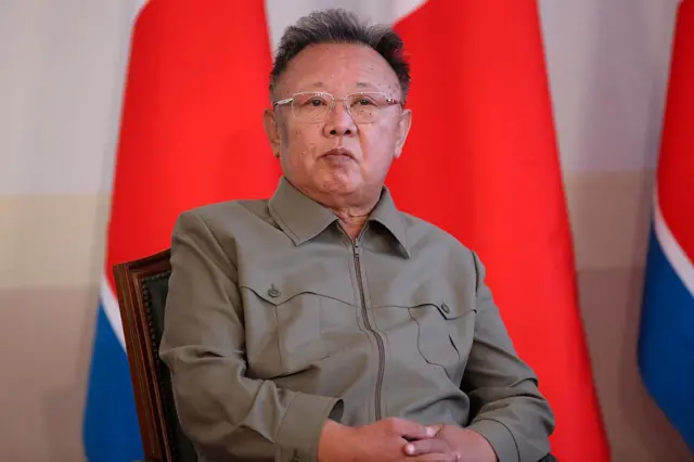 North Korea former leader Kim Jong Il sit on a chair wearing a green shirt and glasses, wit North Korean flags for di background