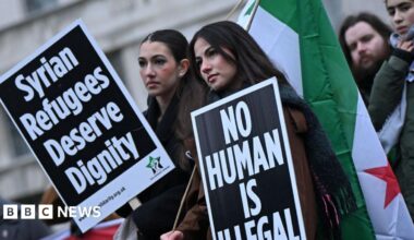 Two female protesters hold Syrian flags and placards reading: "Syrian refugees deserve dignity" and "no human is illegal".