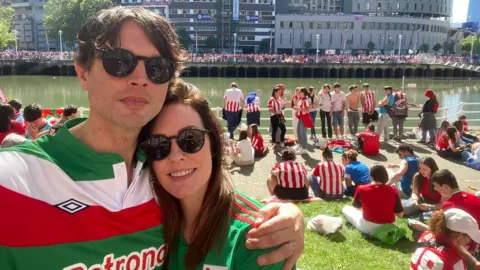 Christopher Evans A man takes a selfies with a woman. He has dark brown hair and sunglasses on and a green, white and red striped football shirt, he has his arm around the woman who has a green shirt on with long dark hair and sunglasses. She smiles at the camera. Behind them there are people in red and white striped shirts sitting and standing by the river. 