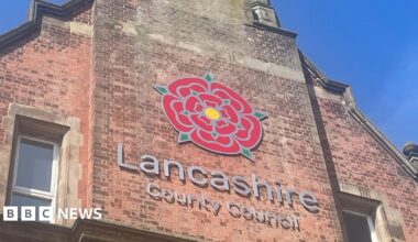 Lancashire County Council sign, with the red rose emblem, on the side of a tall, redbrick building. The photograph was taken on a sunny day with clear blue skies.