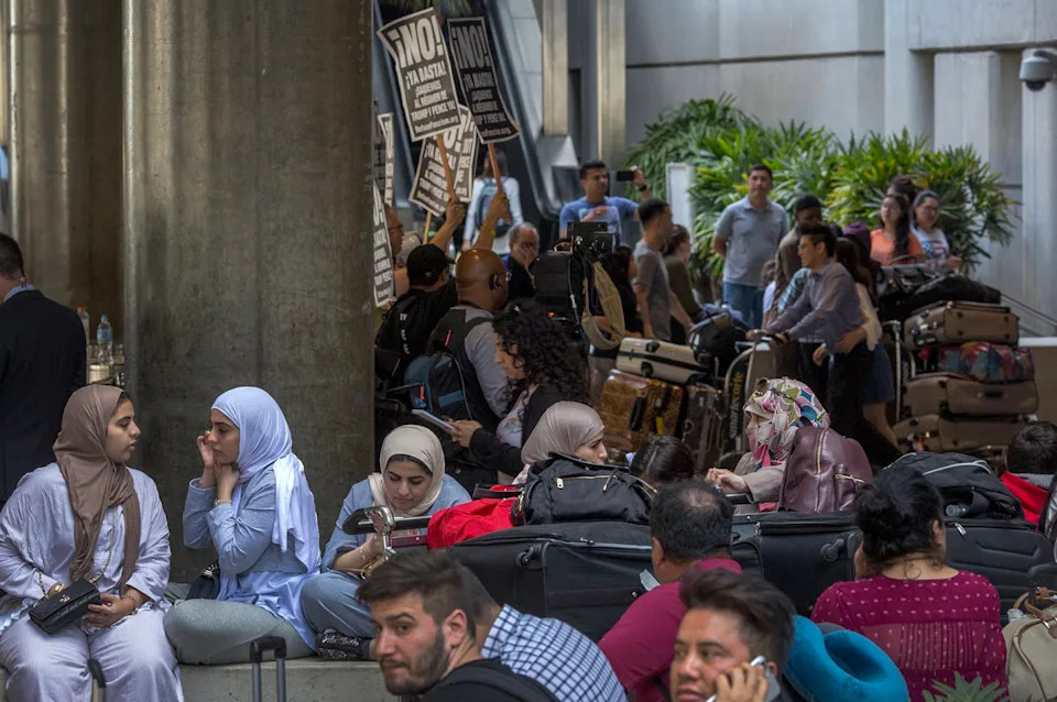 Women and children sit with their baggage in an airport as a protest goes on in the background