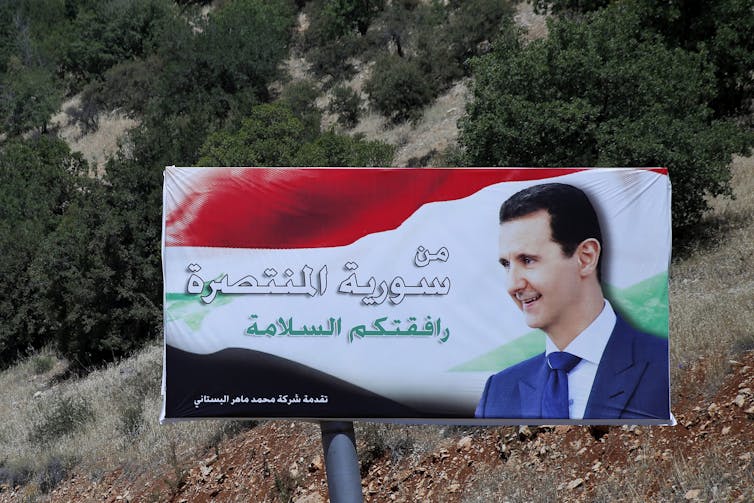 A sign with the face of a smiling dark-haired man reads 'victorious Syria' in Arabic.