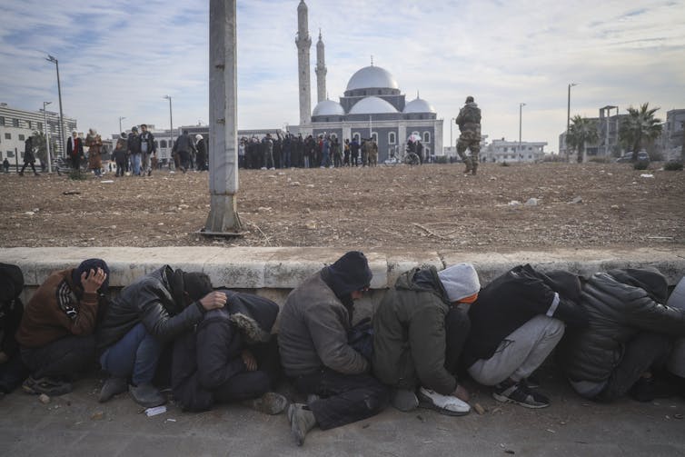 People huddle against a retaining wall and hide their heads as an armed fighter walks nearby