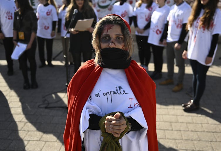 a woman stands in a red cape her hands are bound in front of her and a black cloth covers her mouth. Red paint drips on her forehead.