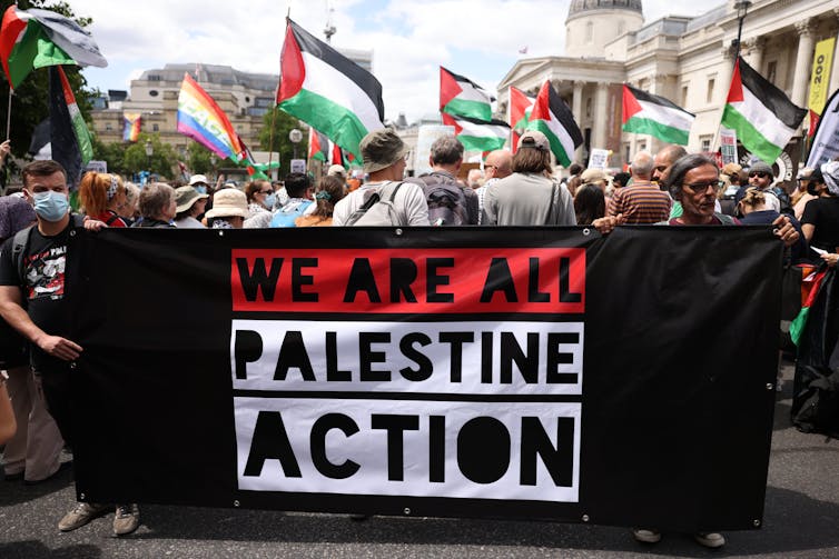 Protestors with a black and red banner which reads 'WE ARE ALL PALESTINE ACTION'.