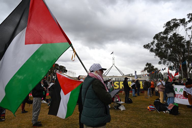 A man carrying a large Palestinian flag among protestors outside Parliament House in Canberra.