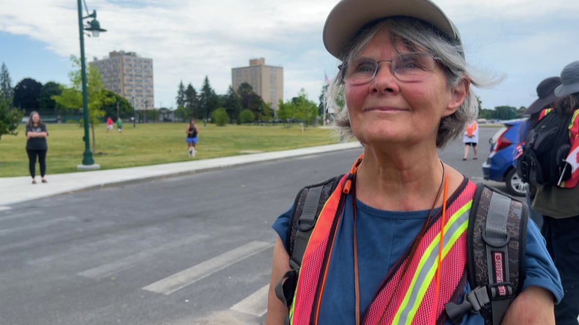 A woman wearing a baseball cap and an orange vest smiles for a photo while standing outside in front of a street.