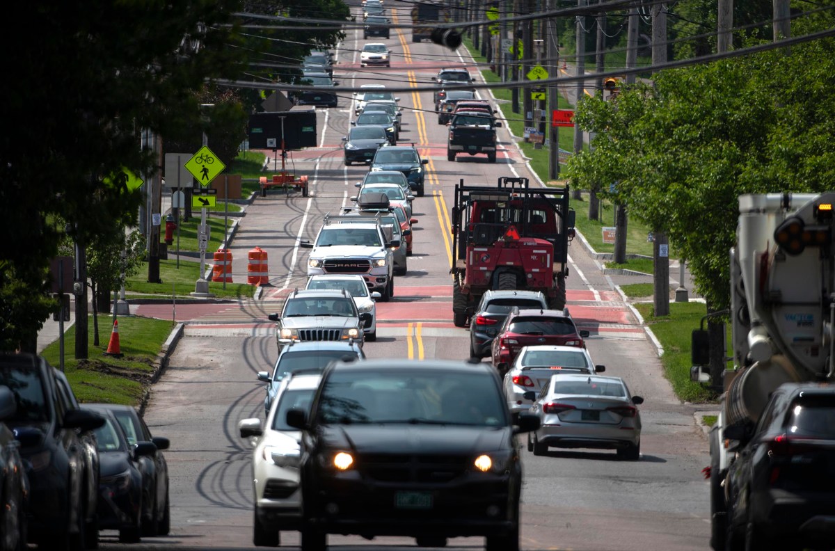 A busy two-lane street with multiple cars and a truck driving in both directions, surrounded by trees and traffic signs on a sunny day.