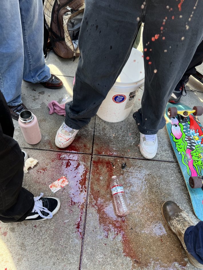 A puddle of blood on the ground. People stand around a young man.