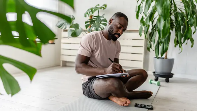 Young African-American man sitting cross-legged on a yoga mat at home, writing in a notebook after a workout, with a smartphone nearby, creating a calm and focused atmosphere
