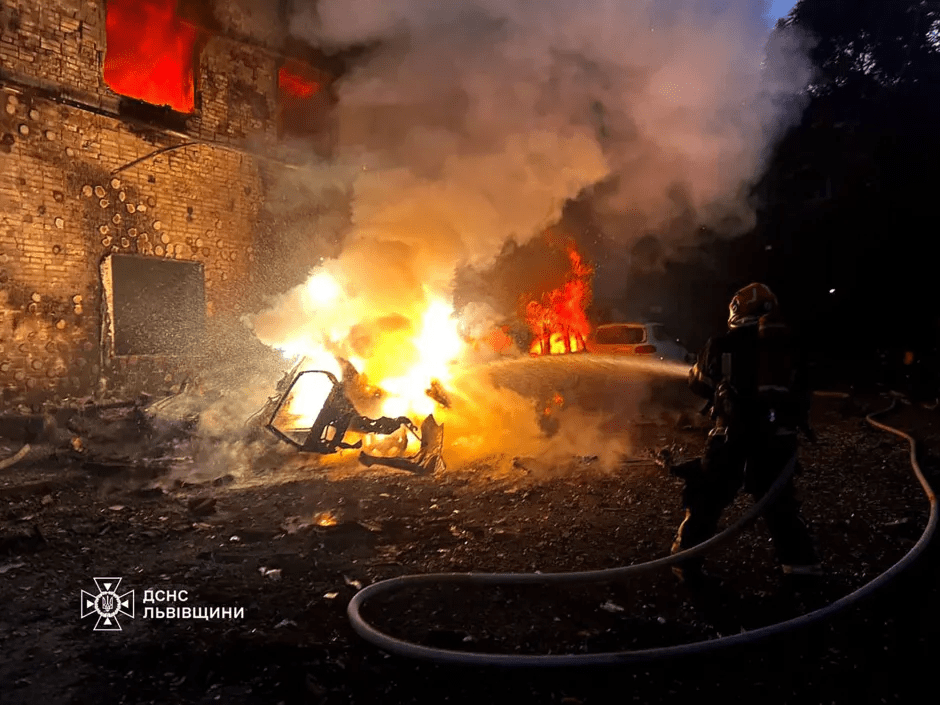 Firefighter battling a large fire engulfing a building and a car.
