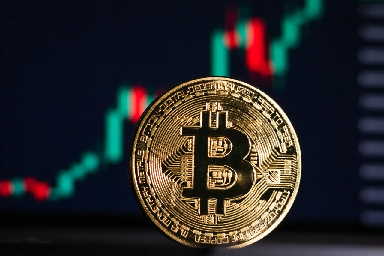 In this photo illustration, a physical Bitcoin (BTC) coin is displayed in front of a screen showing a candlestick chart of the latest Bitcoin price movements.