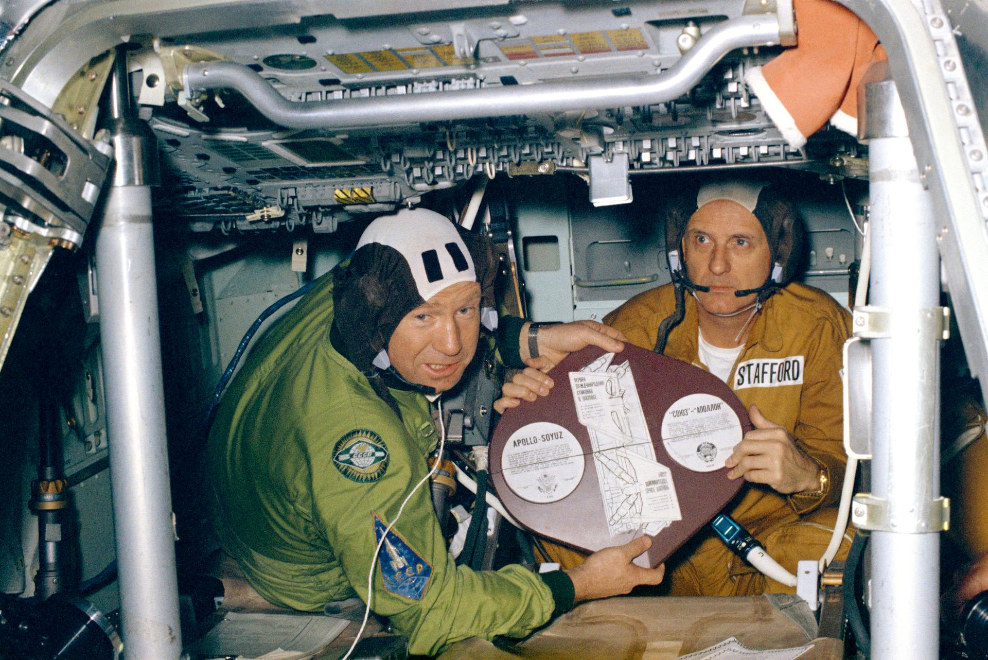 a man in a green flight suit and a man in a orange-tan flight suit hold up a plaque inside a spacecraft
