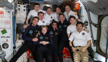 photo of 11 smiling people in a crowded space module. seven are wearing white polos and the other four are wearing dark blue flight suits