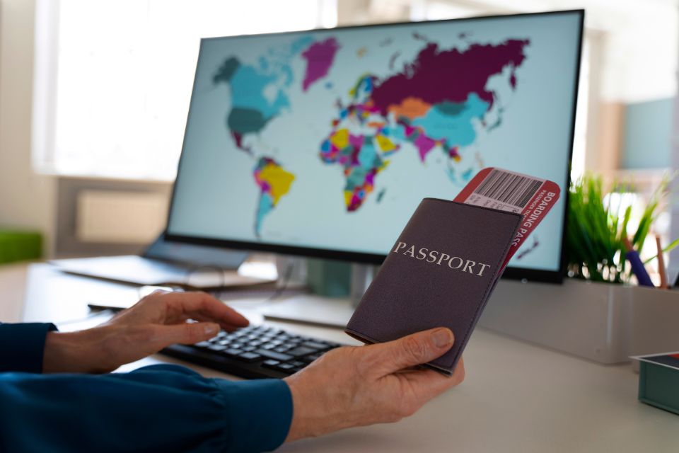 Person holding a passport and boarding pass while using a computer with a colorful world map on the screen.
