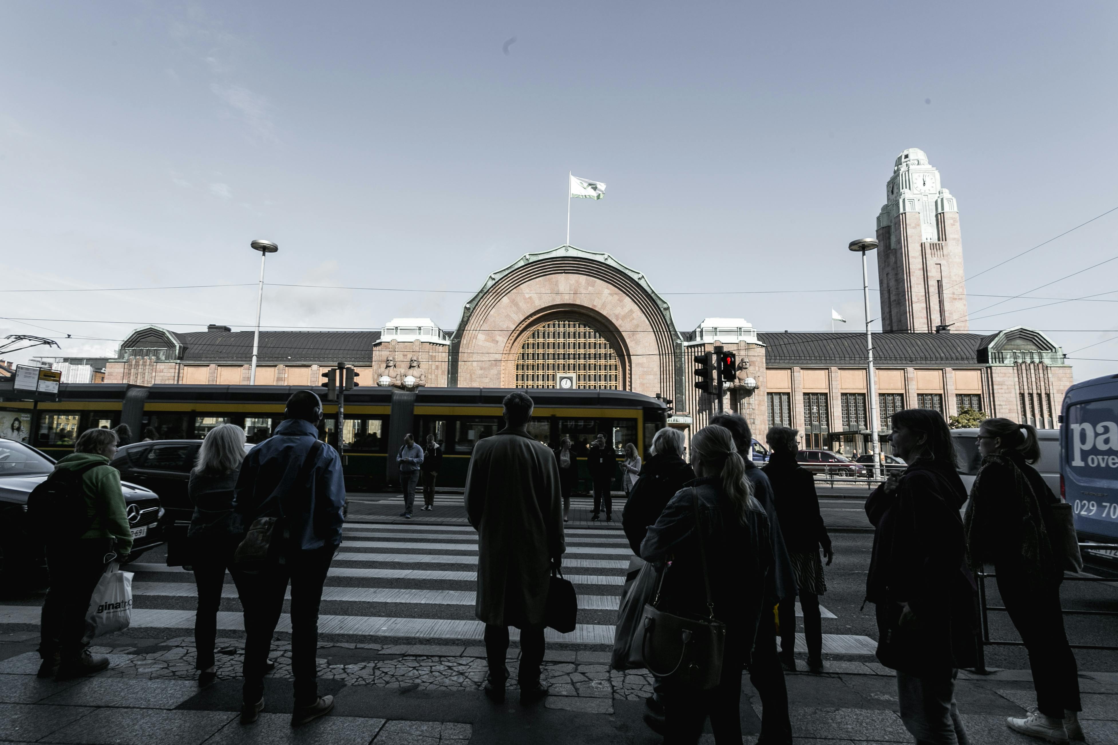 People wait at a crosswalk in front of Helsinki Central Railway Station with a tram passing by and clear skies overhead.
