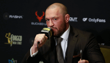 A man in a suit holding a microphone at a press event with sponsor logos in the background.