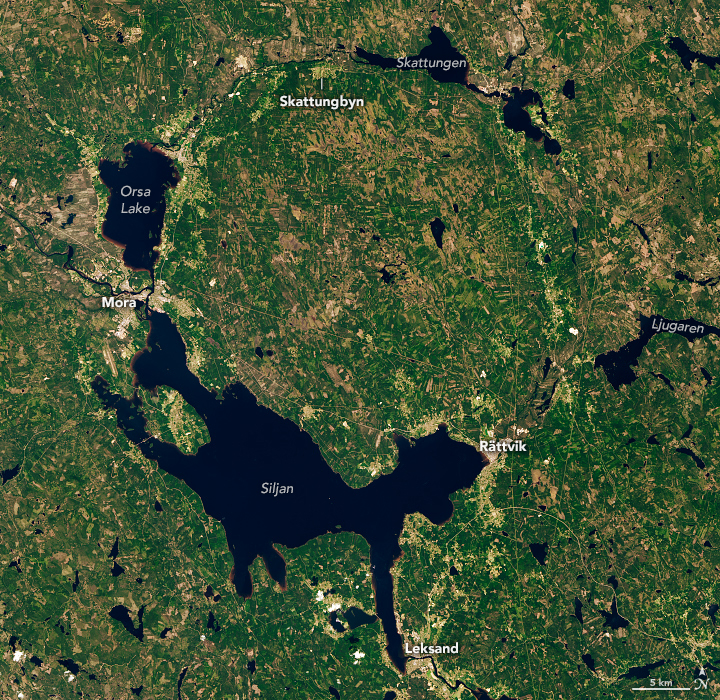 A satellite image shows a ring-like depression in Sweden, remnants of an impact crater, that is partially filled with water. The largest lake, Lake Siljan, is on the crater’s southwest side. Farm fields, towns, roads, and green and brown fields are also visible.