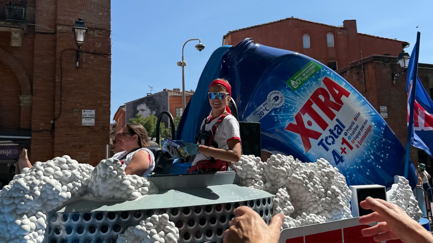 A Tour de France caravan worker in padded getup, wraparound sunglasses, gloves, and a thick headband, distributing free merch from a flat for the X-TRA detergent brand.