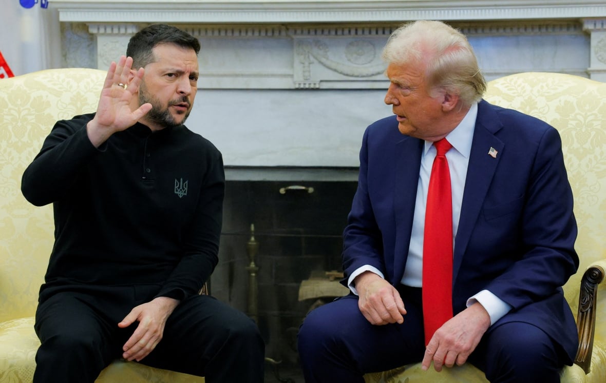 A man in a black suit raises his hand as another man in a blue suit looks at him in anger.