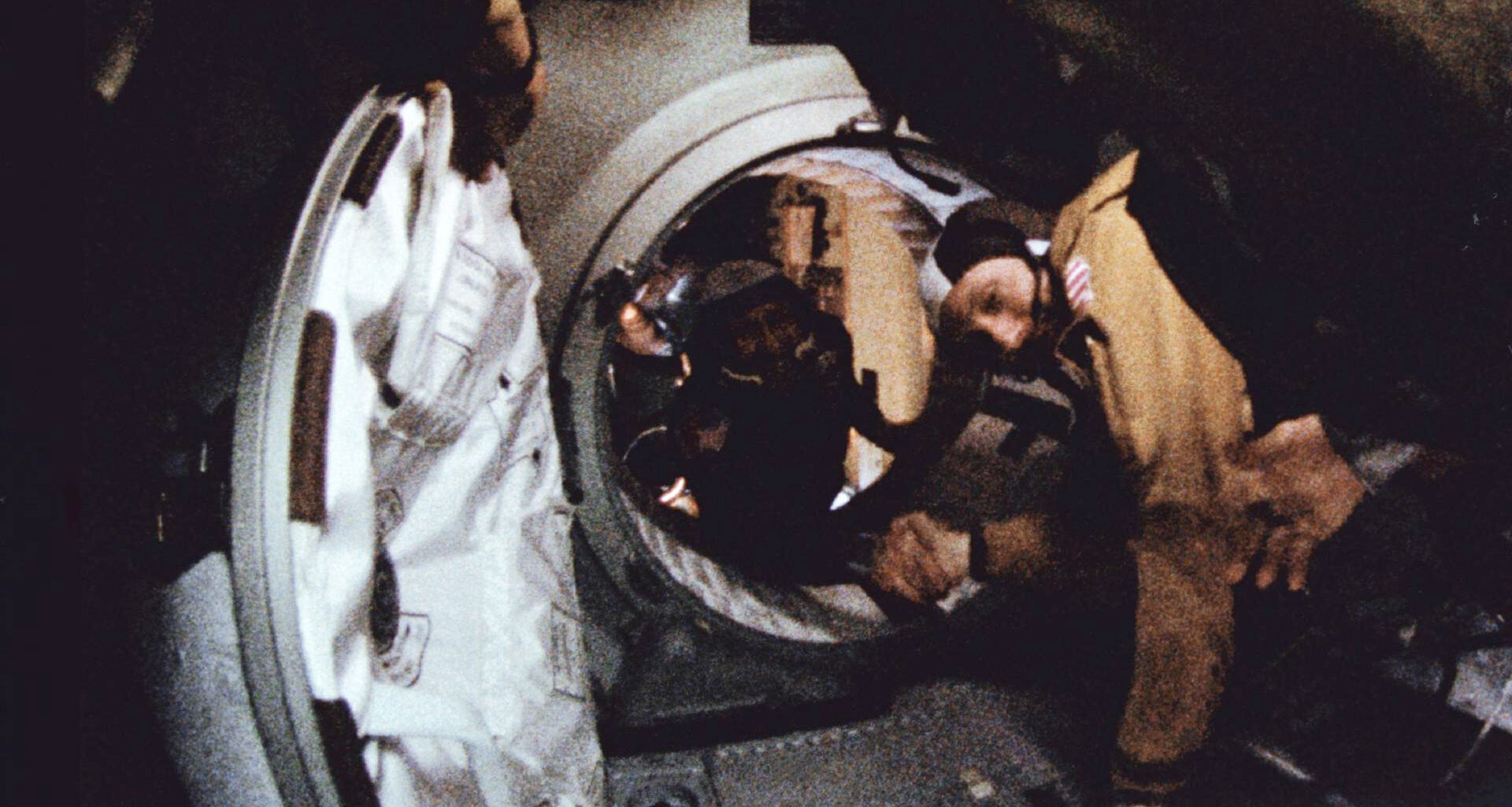 two men floating in space reach out and shake hands across the open threshold of a spacecraft hatchway