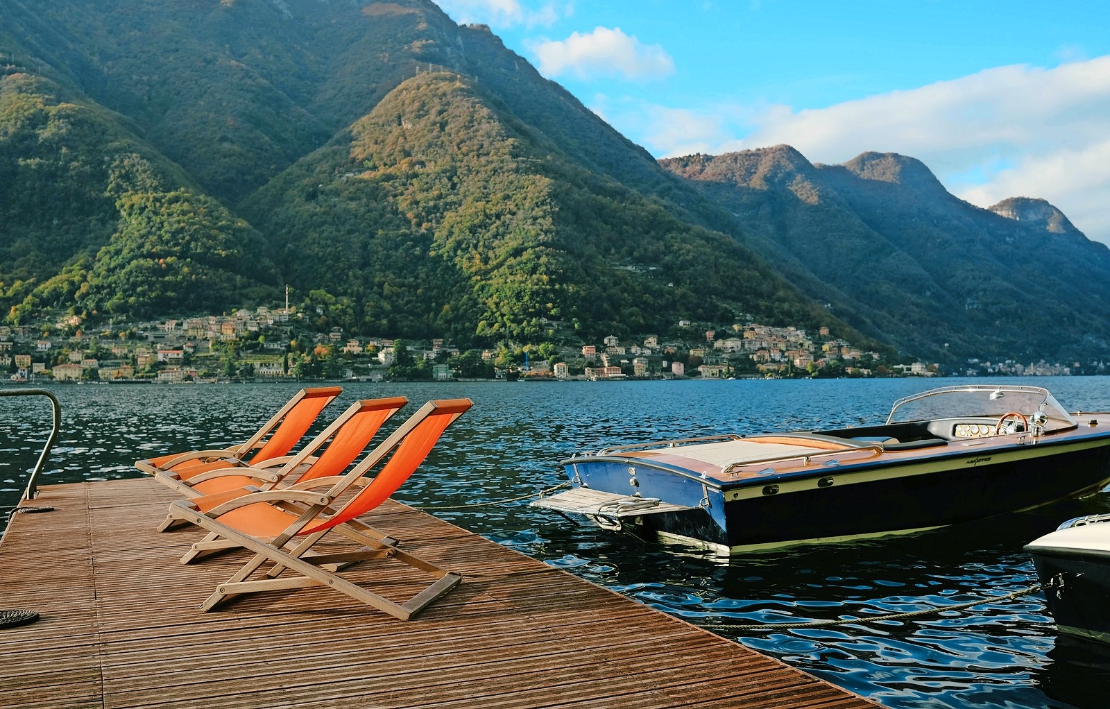 Three lounge chairs on a dock by a lake with a boat and mountains in the background.