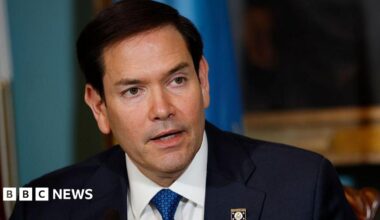 Marco Rubio man wearing a blue tie, dark suit and white shirt looks off to the right. His mouth is slightly open as he is mid-conversation. He is wearing a pin with the American eagle on his right lapel.