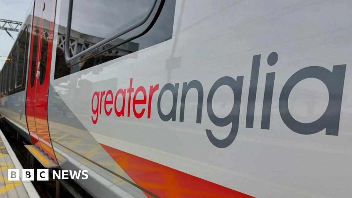 A Greater Anglia train with the company name on the side in red and grey lettering. The train is painted in the red, white and grey livery of the company. Black-framed windows and red doors can be seen. The train is alongside a platform paved with small grey squares.