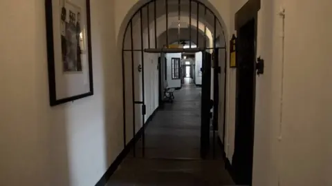 A prison corridor an arched ceiling and white walls on either side. There is a black gate door, which is open, and on the left is a black and white picture hanging on the wall. 
