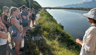 A group of students stand on a grassy path looking at a river. An instructor stands in the foreground.
