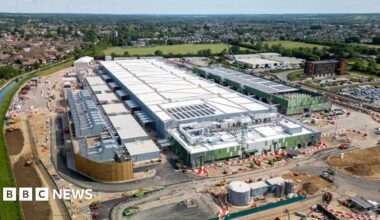 A large white data centre building under construction in Hertfordshire, surrounded by green land, a river and housing estates further afield.