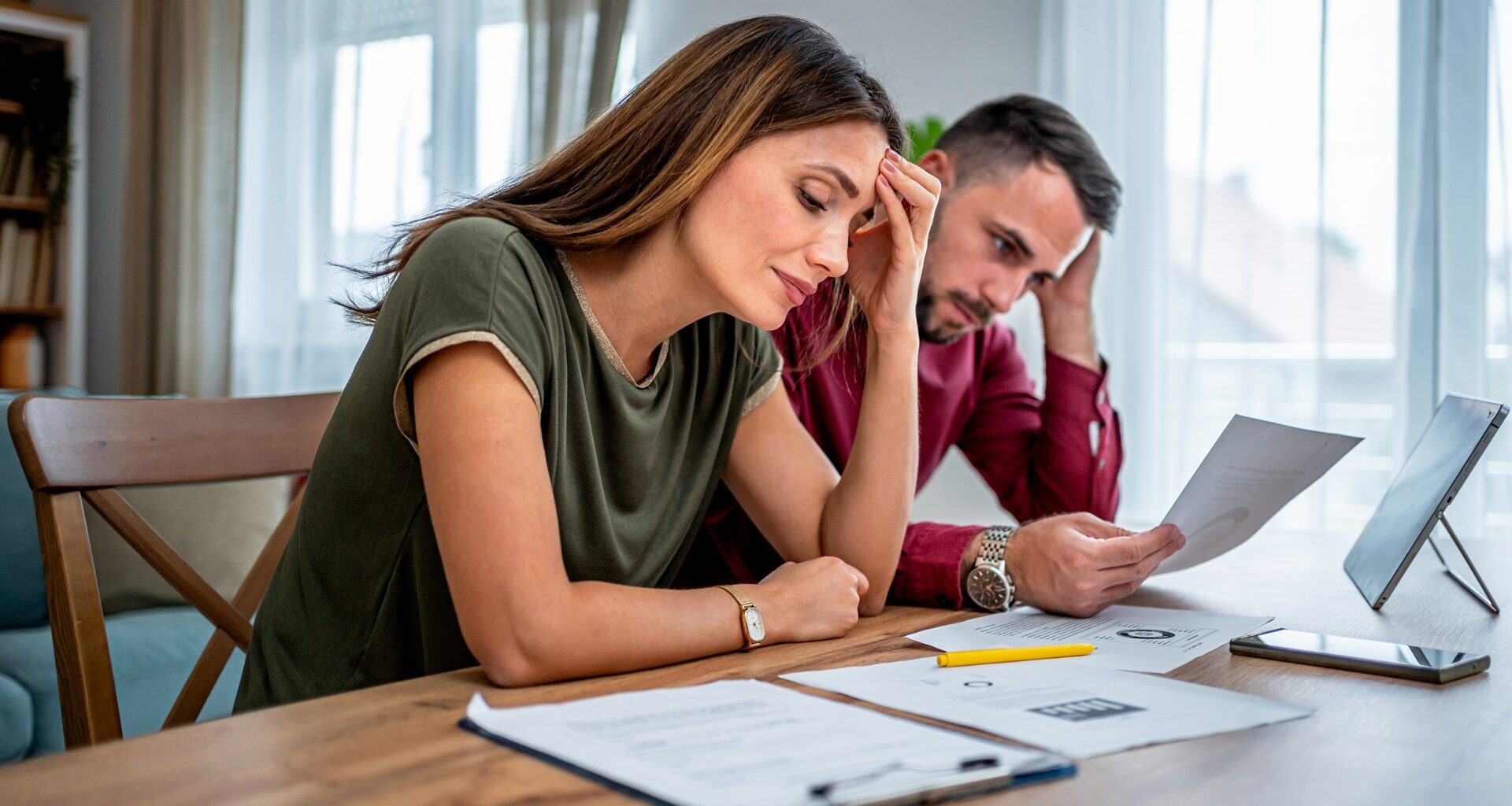 A young couple with worried expressions sits at a dining table, reviewing financial paperwork with a tablet and phone.