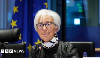 Head of the European Central Bank, Christine Lagarde, sitting wearing a black leather jacket and a black and white scarf around her neck, with the EU flag behind her