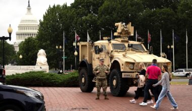 A person in a National Guard uniform stands next to a tan military vehicle parked on a red brick pavement. Three people walk in front of the military vehicle. The U.S. Capitol Dome and various trees can be seen in the background.