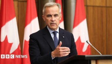 Canada's Prime Minister Mark Carney speaking at a podium and gesturing with his right hand. He is wearing a black suit with a navy blue patterned tie and a light blue button-up shirt. Behind him is a row of Canadian flags on poles.
